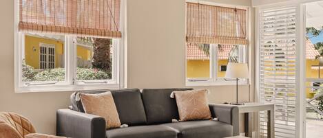Una sala de estar con un sofá, sillones, una mesa de centro y una lámpara. Hay ventanas con persianas y vistas al exterior.