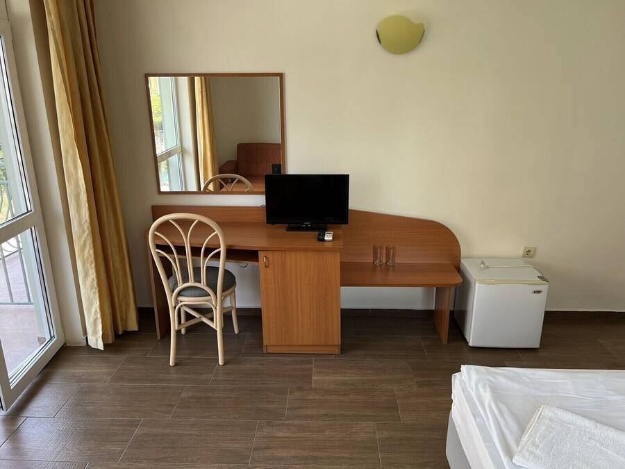 Habitación de hotel con escritorio de madera, una silla, televisor, refrigerador pequeño y cama con manta blanca.