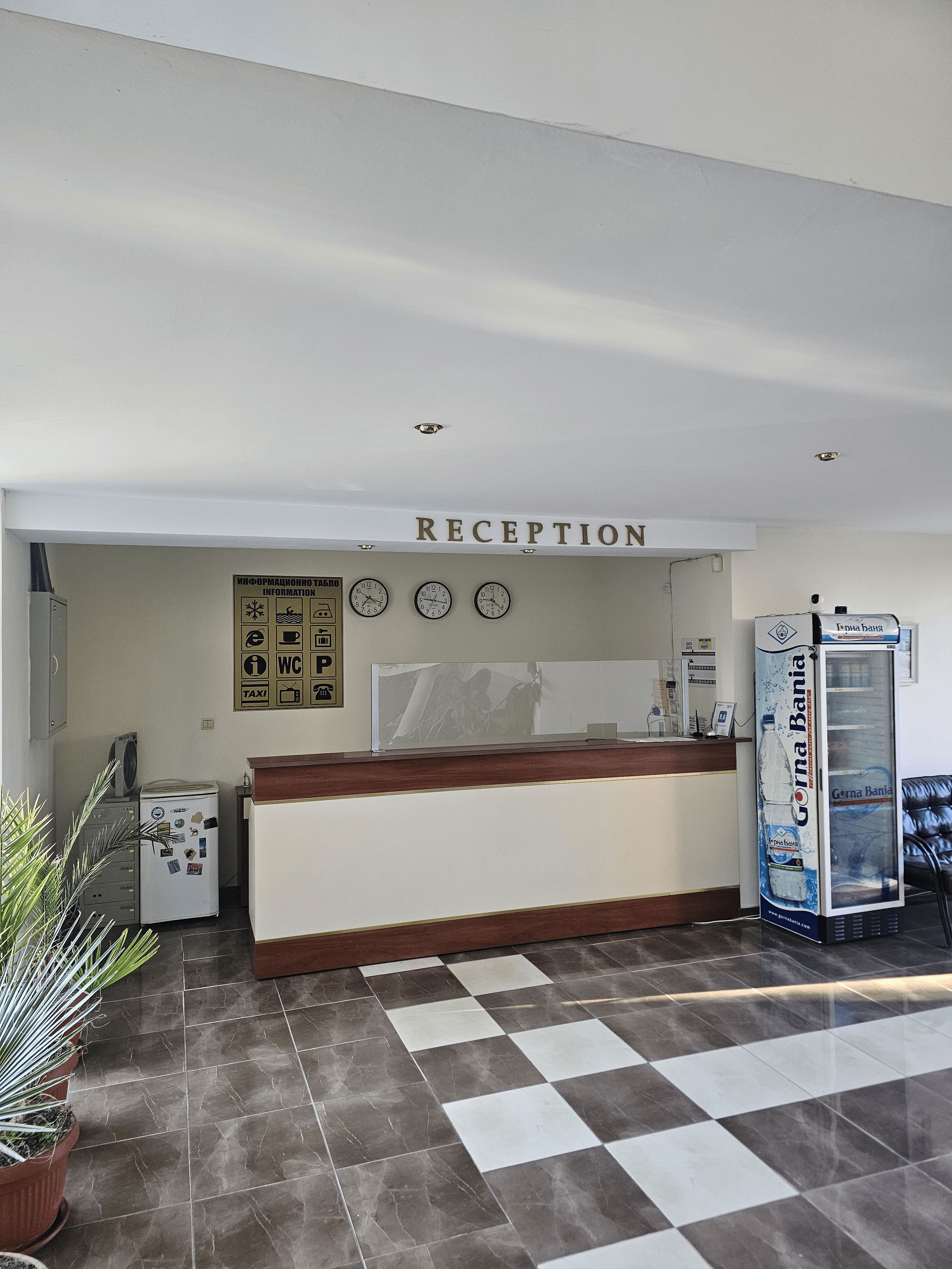 Zona de recepción con mostrador, un panel informativo en la pared, refrigerador, máquina expendedora y suelo a cuadros.