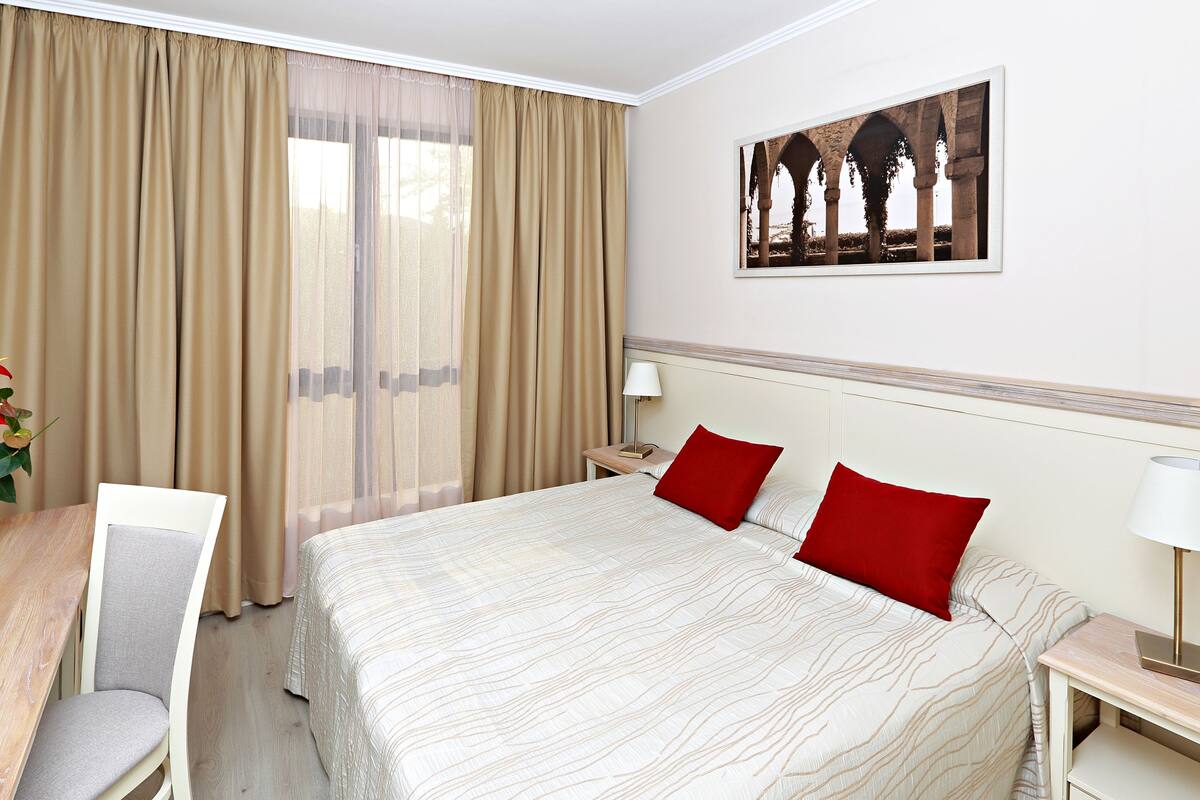 A hotel room with a large bed, two red pillows, a wooden desk with a chair, a nightstand with a lamp, and a framed picture on the wall.