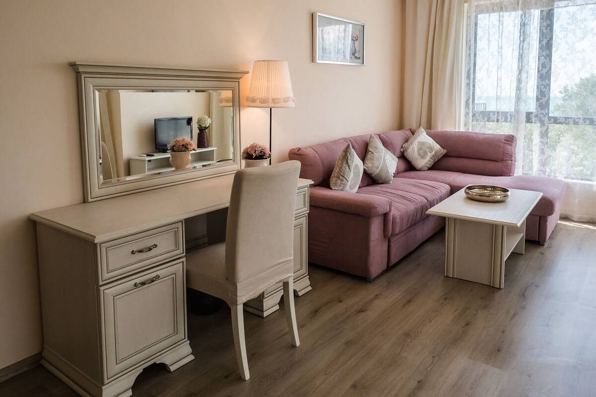 A room with a desk, a pink sofa, a chair, a lamp, and a window with curtains.