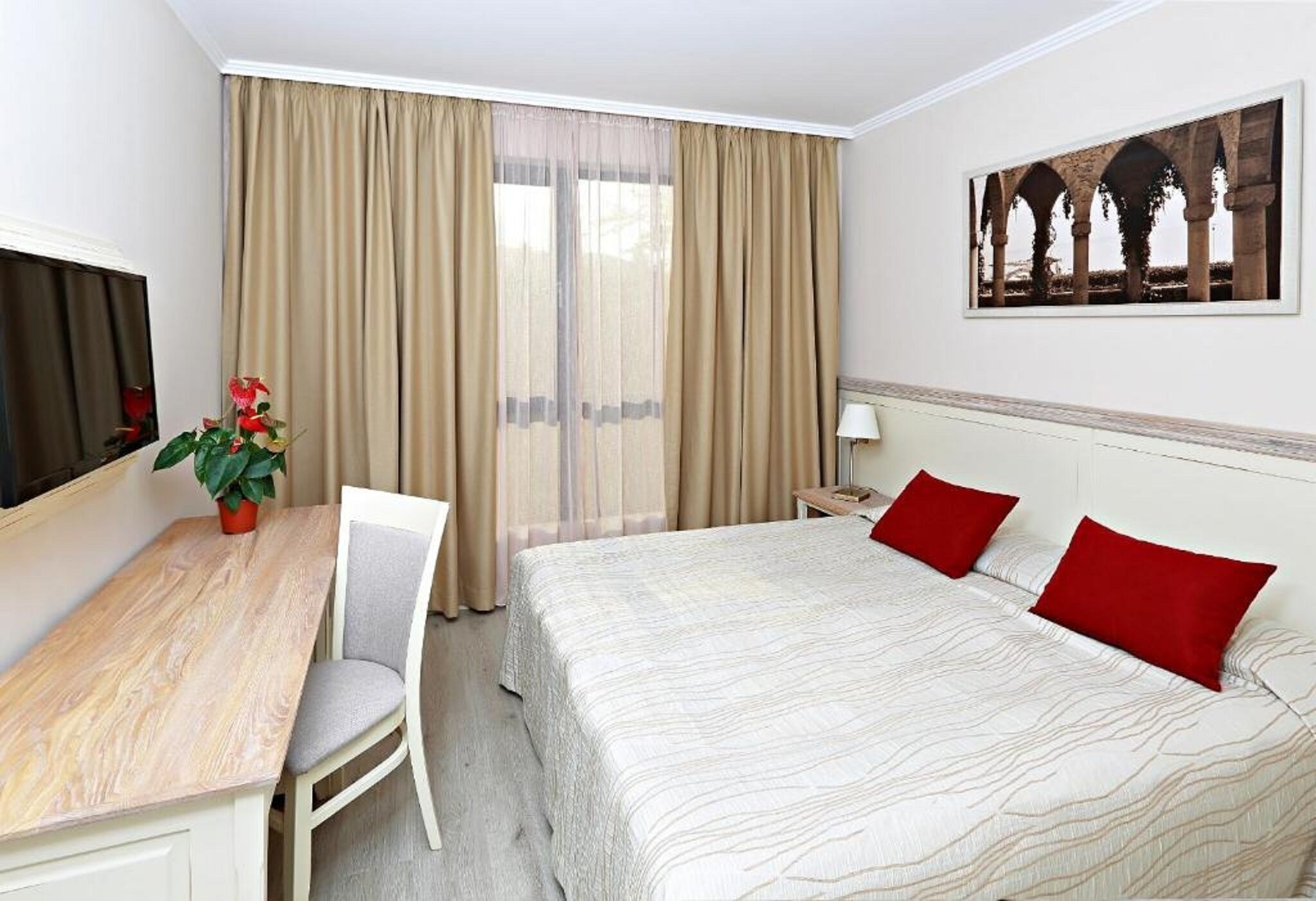 A hotel room with a bed, a desk, a chair, a TV, a lamp, and a painting on the wall.