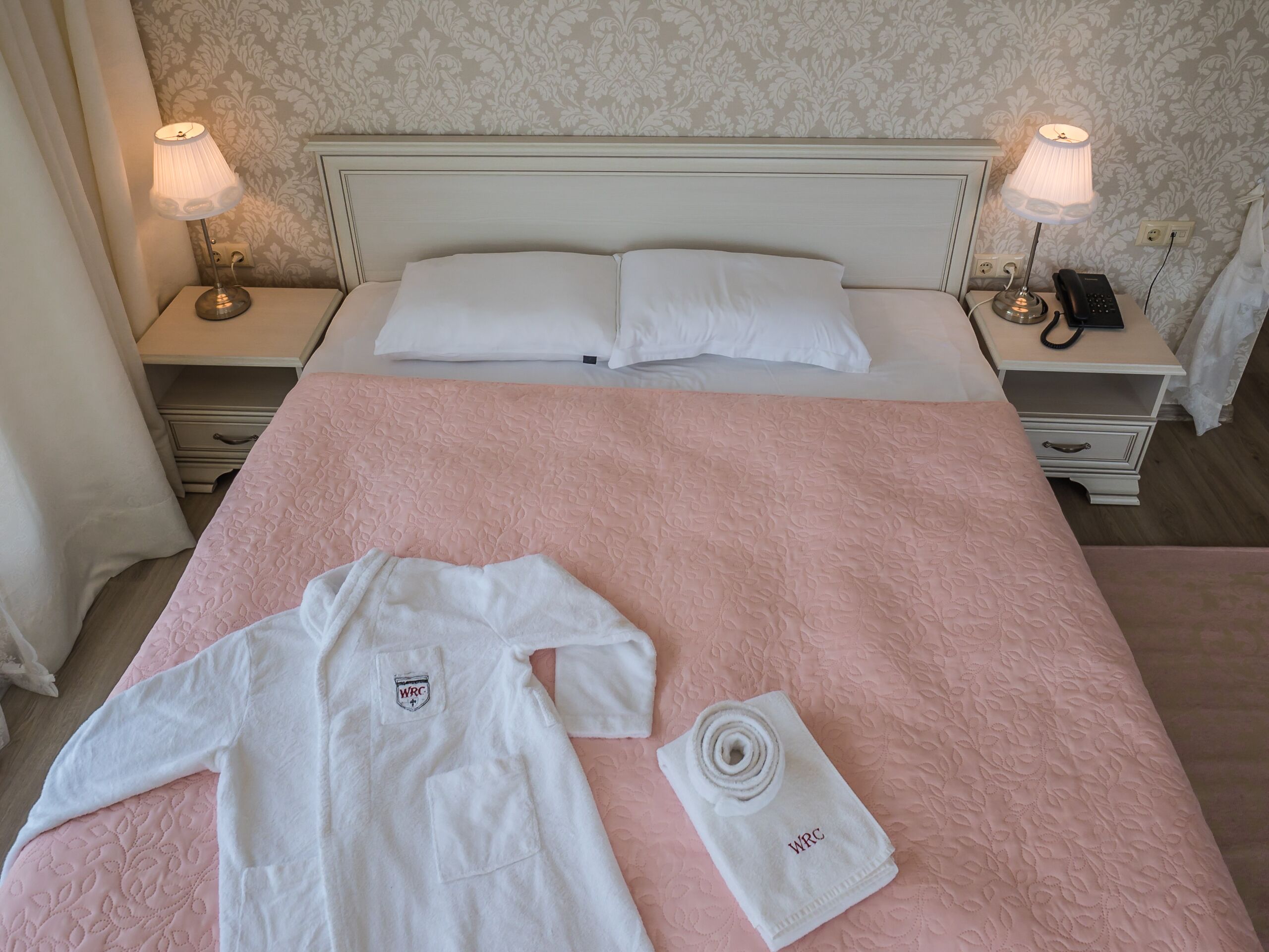 Una cama bien hecha con un edredón rosa, dos almohadas blancas y una bata y toalla blancas sobre la cama.