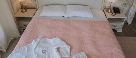 Una cama bien hecha con un edredón rosa, dos almohadas blancas y una bata y toalla blancas sobre la cama.