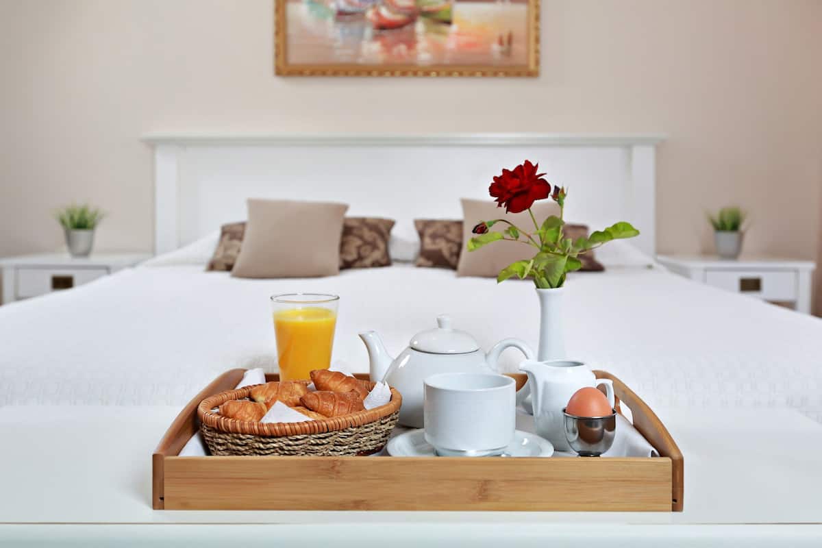A neatly made breakfast tray with croissants, a glass of orange juice, a teapot, a cup, and a rose in a vase on a bed.