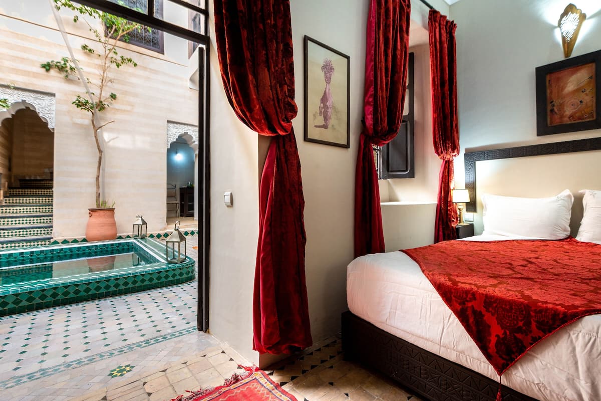 Une chambre avec un lit, un rideau rouge, un tableau encadré et une vue sur l’espace piscine.
