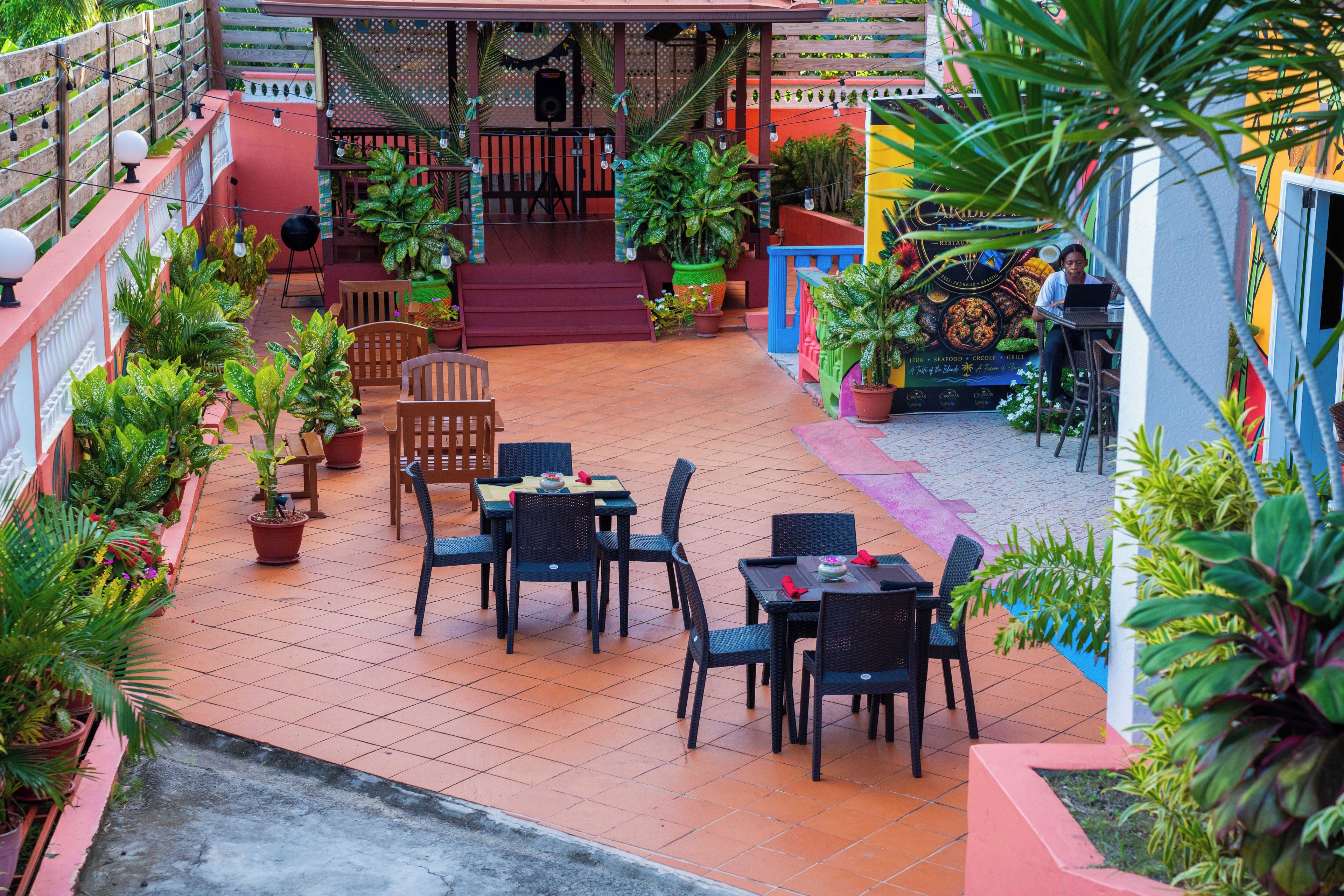 Un espace extérieur aménagé en terrasse, avec plusieurs tables et chaises, entouré de plantes en pot et d’une clôture décorative.