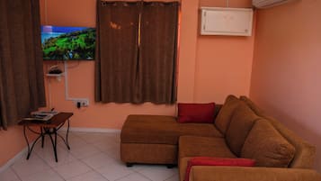 A living room with a brown sofa, a red pillow, a television on the wall, and an air conditioning unit.
