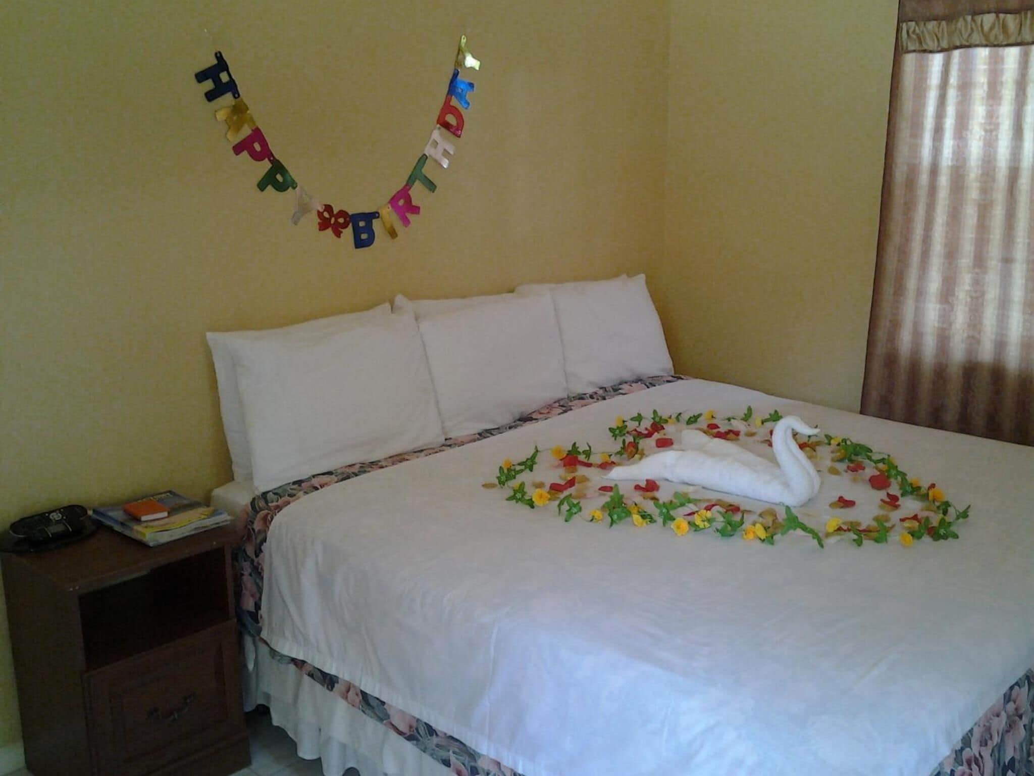 A neatly made bed with a swan-shaped decoration made of flowers and leaves, a 'Happy Birthday' banner, and a bedside table with a book and a phone.