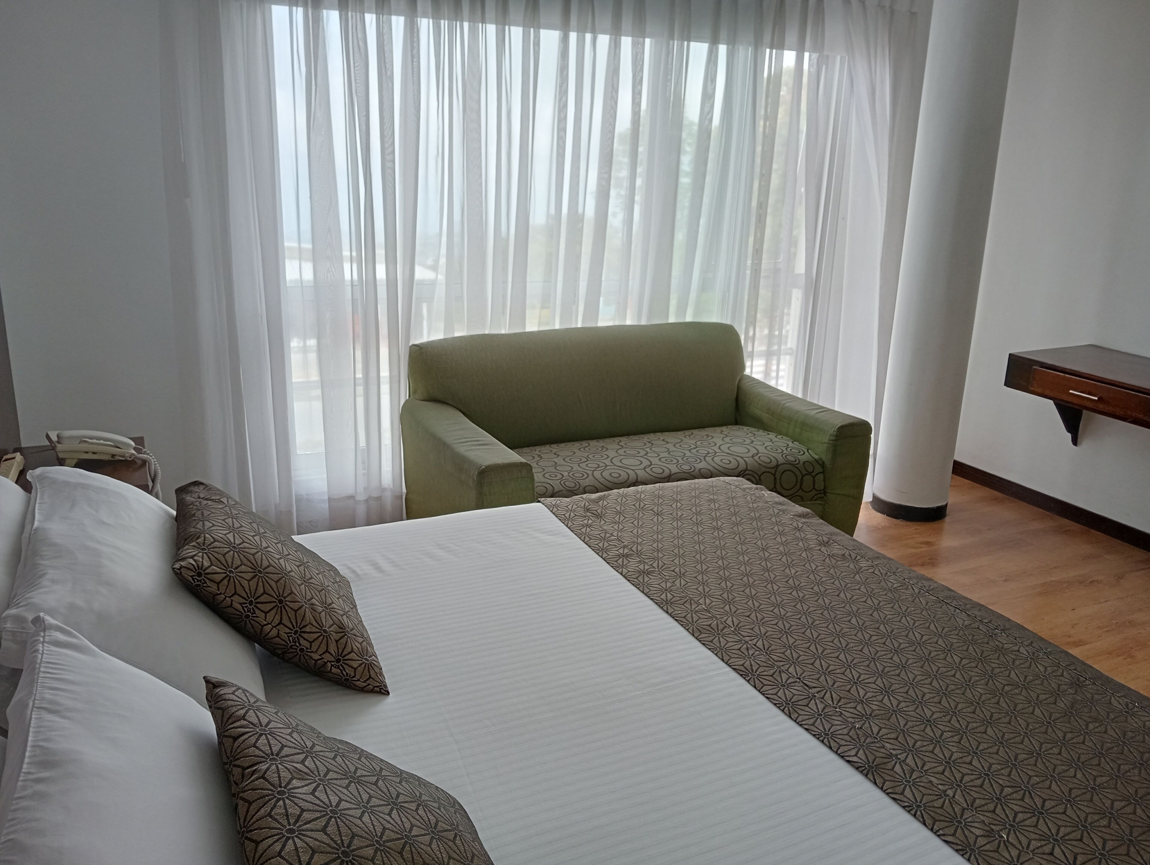 Ein Hotelzimmer mit einem Bett, einem grünen Sessel, einem Nachttisch und einem Fenster mit durchsichtigen Vorhängen.