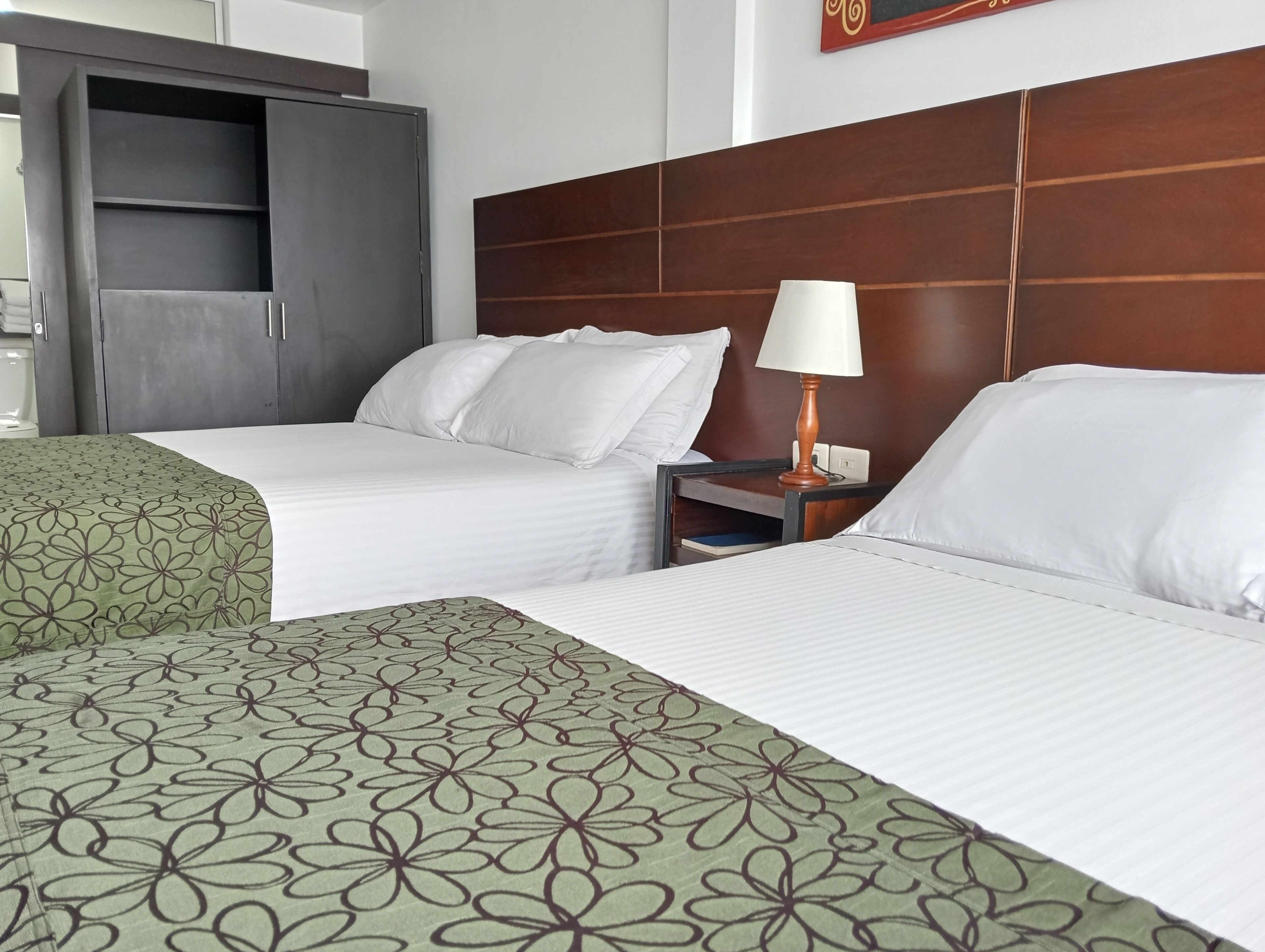 Habitación de hotel con dos camas, un armario, una mesita de noche con lámpara y un cuadro colgado en la pared.