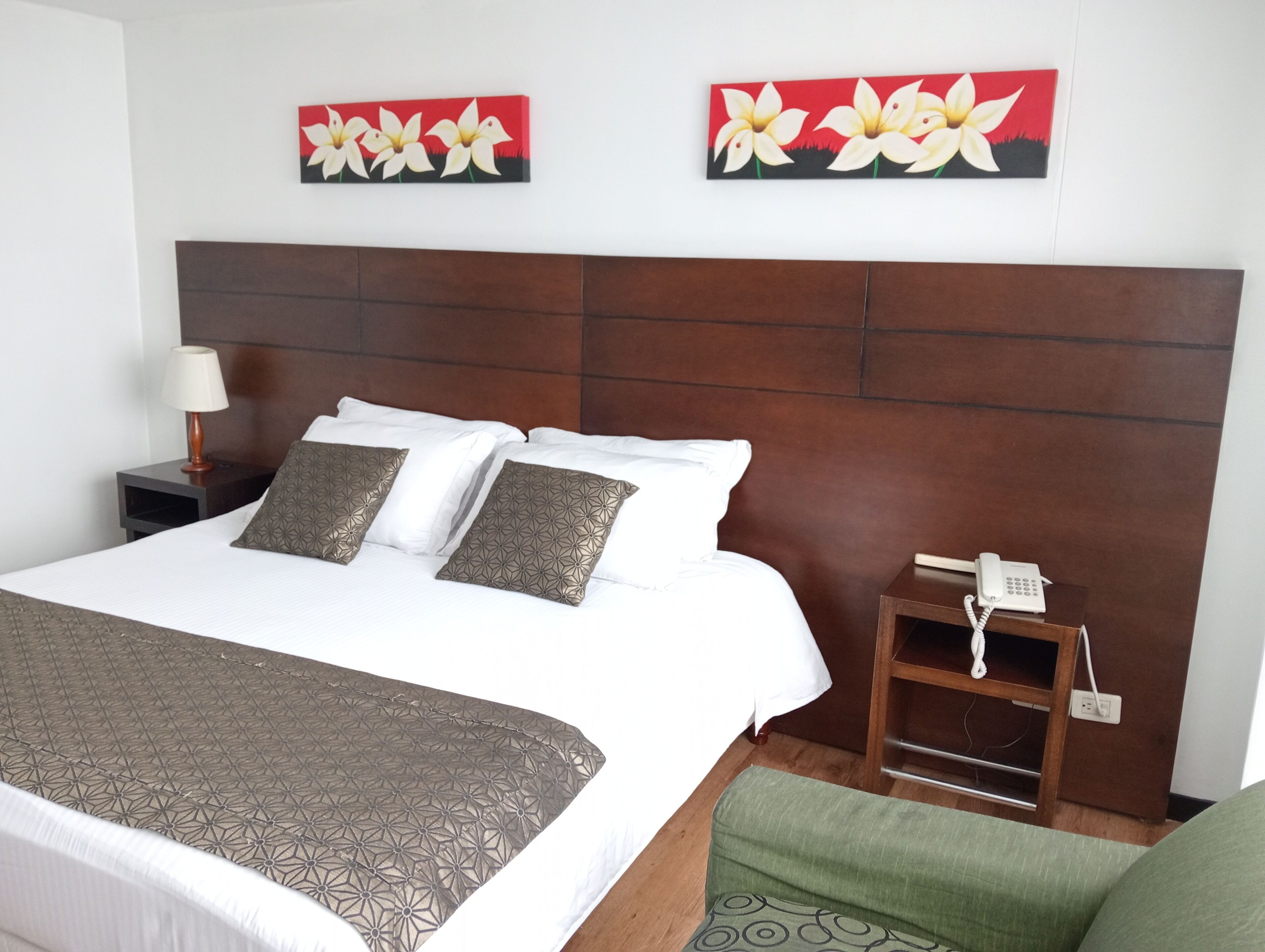A hotel room with a large bed, two pillows, a bedside table with a lamp, a phone, and two wall-mounted paintings.