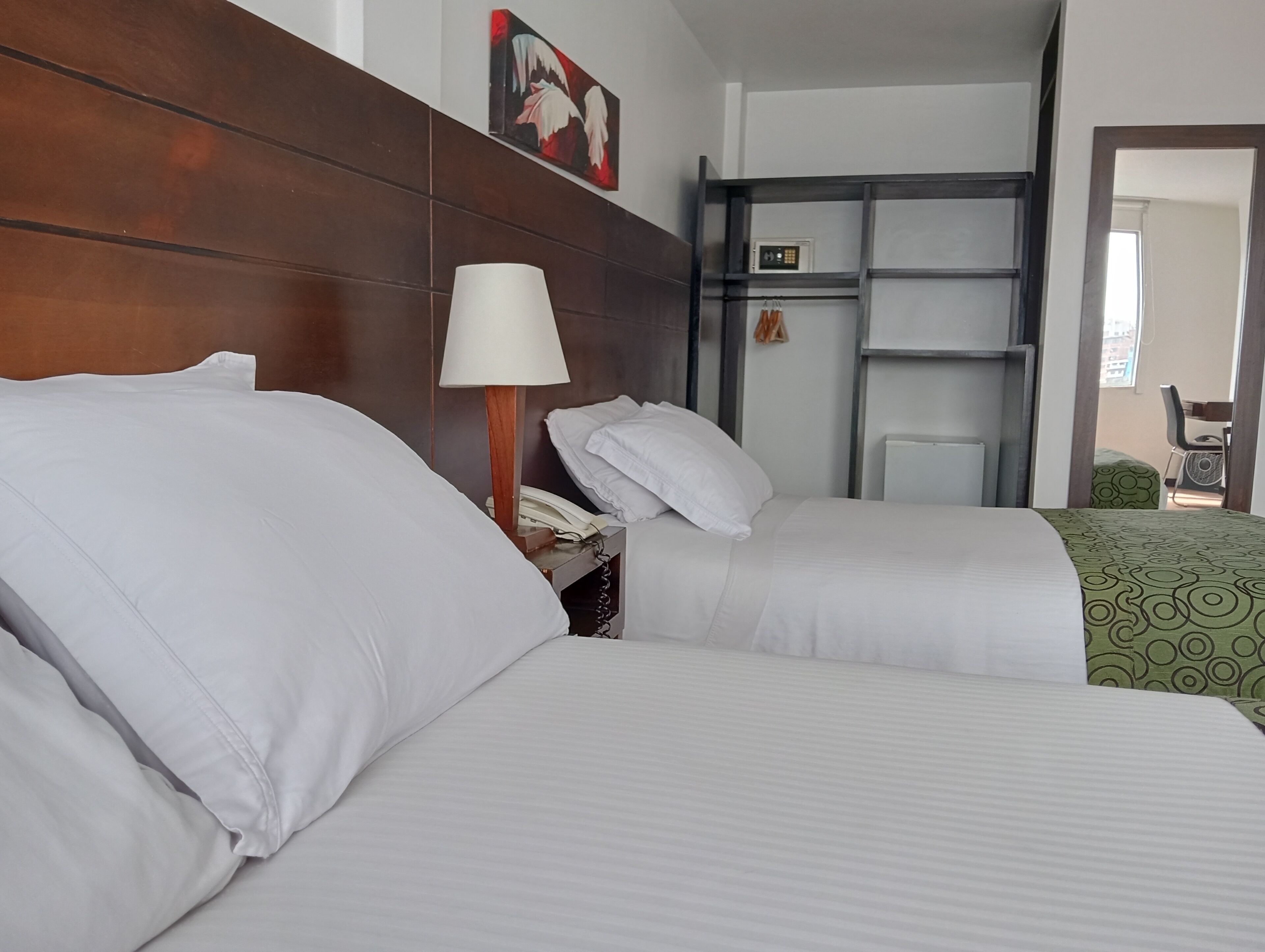 A hotel room with two beds, a bedside lamp, a telephone, and a small shelf with a clock.