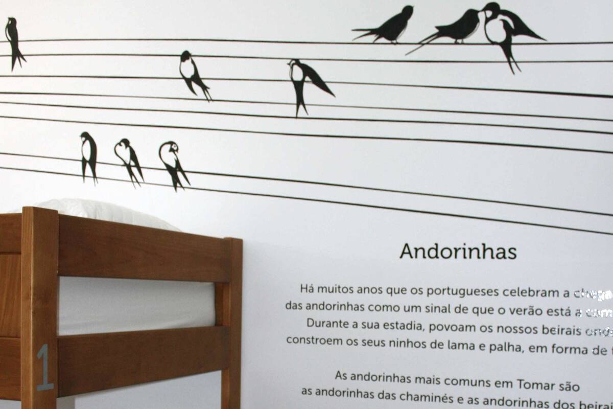 A bunk bed with a wooden frame and white bedding, a wall with a black and white mural of birds, and a text panel titled "Andorinhas" with information about Portuguese traditions.