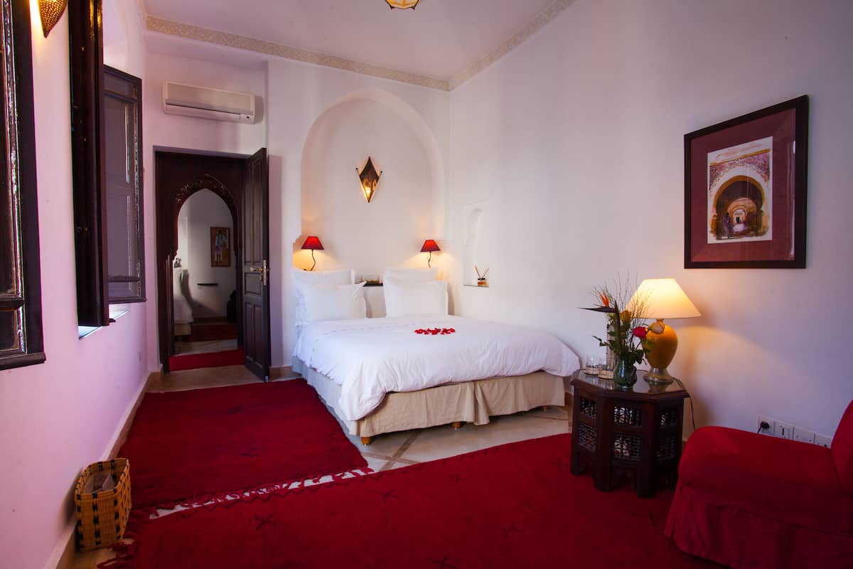 Una habitación de hotel con una cama grande, dos lámparas de noche, una alfombra roja, una mesita con un florero y un cuadro en la pared.