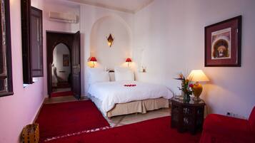 Een hotelkamer met een groot bed, twee bedlampen, een rood vloerkleed, een kleine tafel met een vaas en een ingelijst schilderij aan de muur.