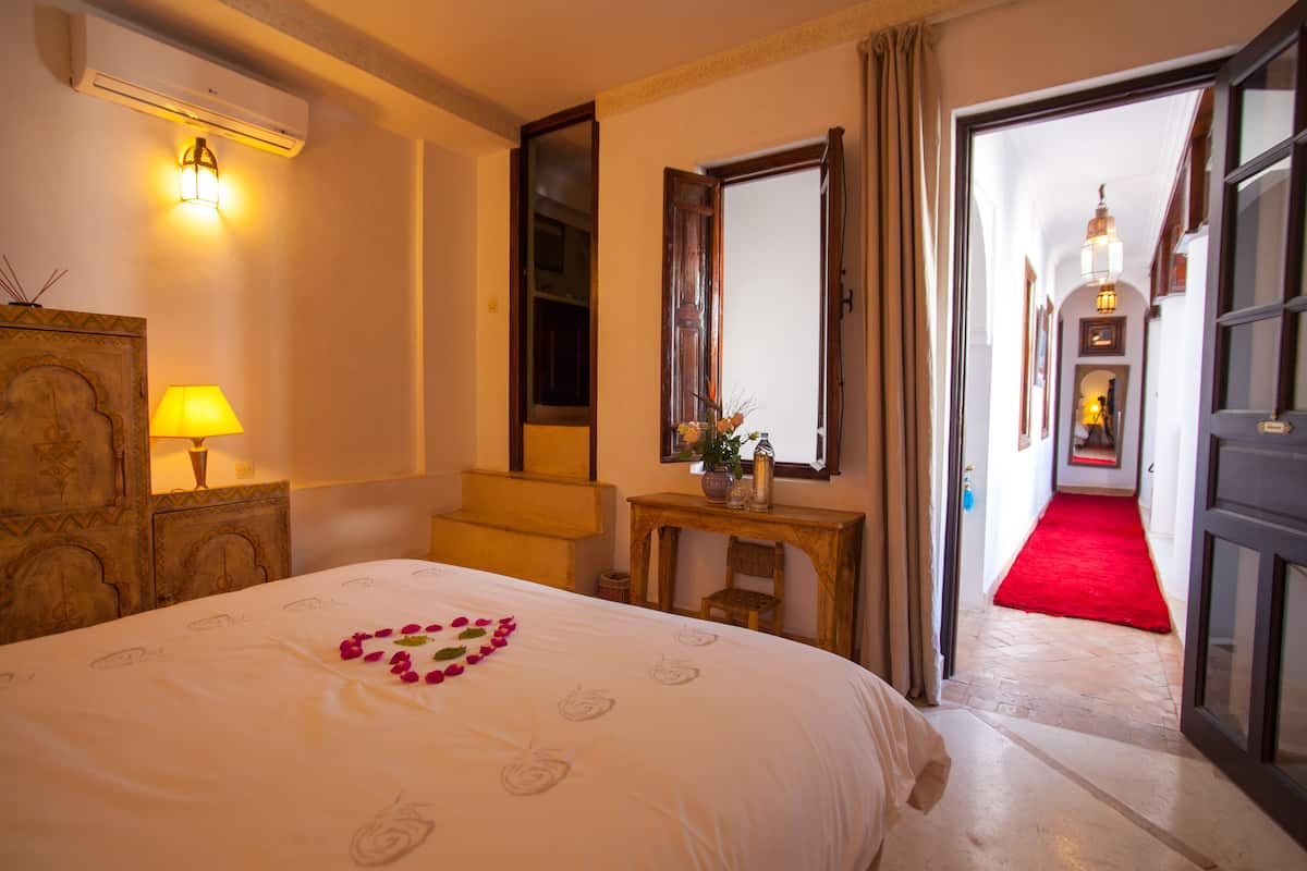Una habitación de hotel con una cama, una mesita de noche con lámpara, un aparador de madera, un espejo, un pasillo con alfombra roja y una puerta que da a otra habitación.