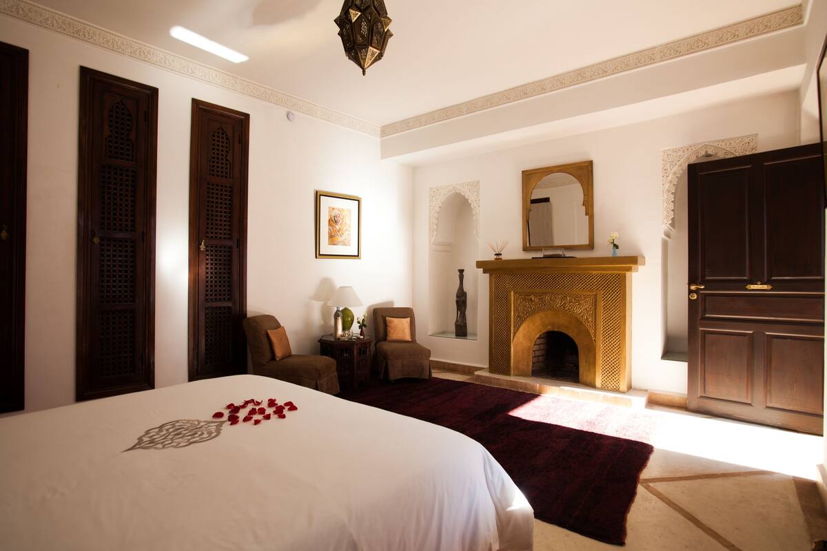 Habitación de hotel con una cama grande, una chimenea, dos sillones, una mesita con una lámpara y un espejo grande sobre la chimenea.