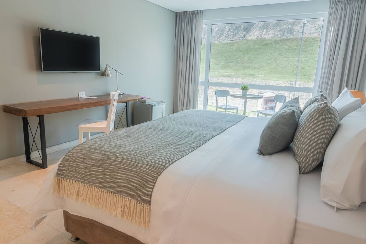 Una habitación de hotel moderna con una cama grande, un escritorio, una silla, un televisor y un balcón con vistas a las colinas.