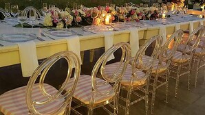 An outdoor dining setup with clear chairs and a long table, set against a backdrop of a rocky hill and city buildings. - GHL San Lazaro Art Hotel (Cartagena)
