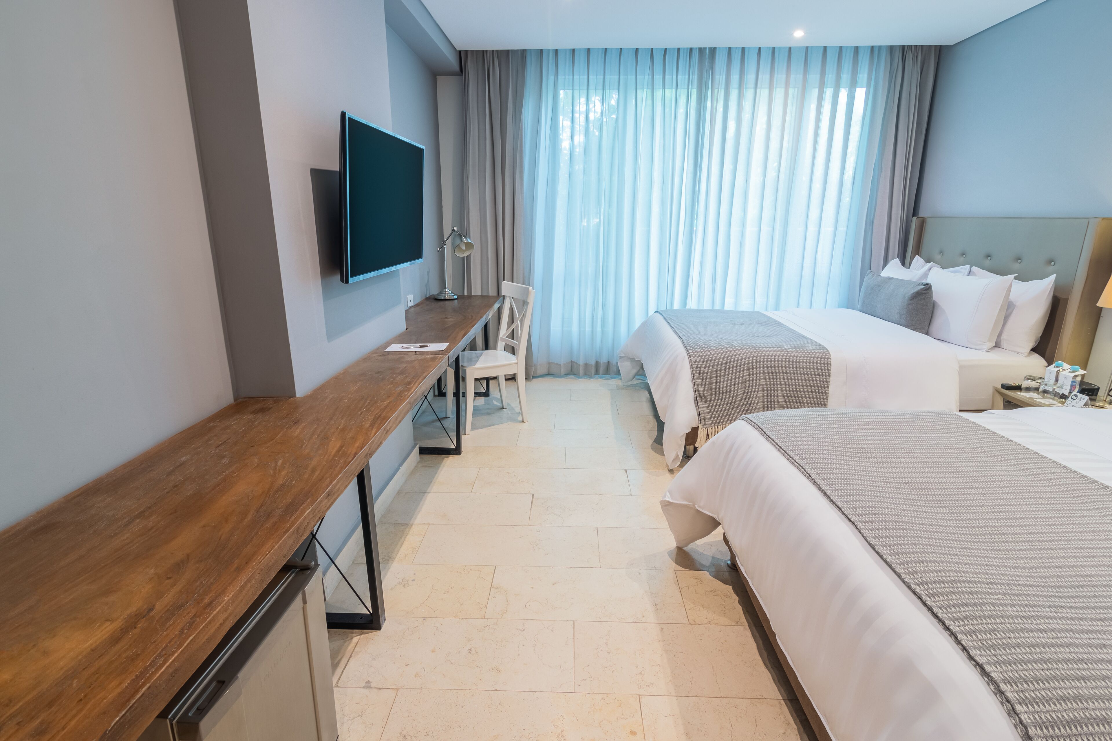 Une chambre d’hôtel avec un bureau en bois, un téléviseur fixé au mur, deux lits avec du linge de lit blanc, une chaise et une grande fenêtre avec des rideaux légers.