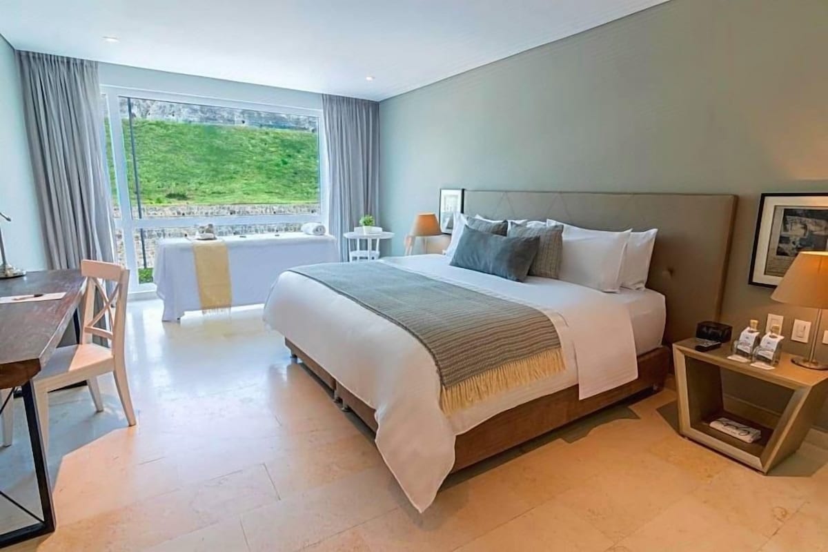 A modern hotel room with a large bed, a desk, and a view of a green landscape through the window.