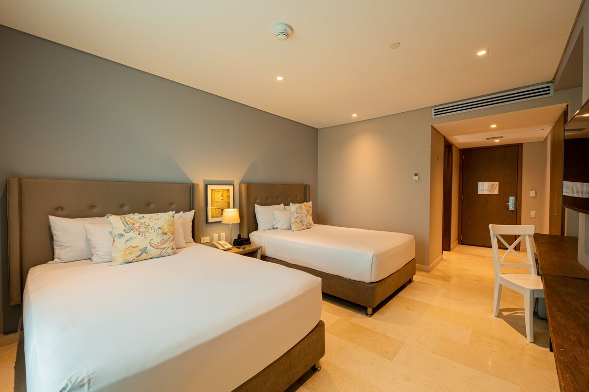 A hotel room with two beds, a desk, a chair, a nightstand, and a door leading to another room.