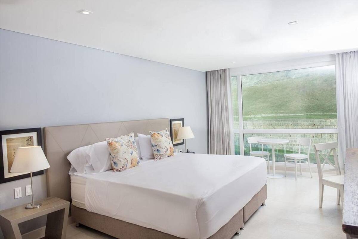 Habitación de hotel con una cama grande, dos mesitas de noche, un ventanal con cortinas y un balcón con sillas.