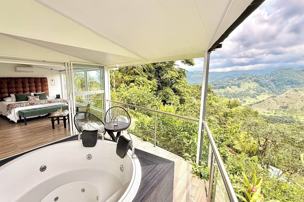 A bedroom with a large bed, a balcony with a hot tub, and a scenic view of a valley.