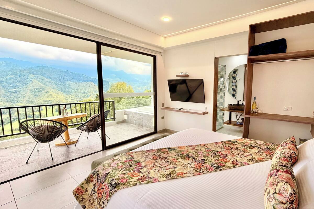 A hotel room with a large bed, a TV mounted on the wall, a balcony with chairs, and a view of mountains.