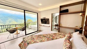 Una habitación de hotel con una cama grande, un televisor montado en la pared, un balcón con sillas y vistas a las montañas.