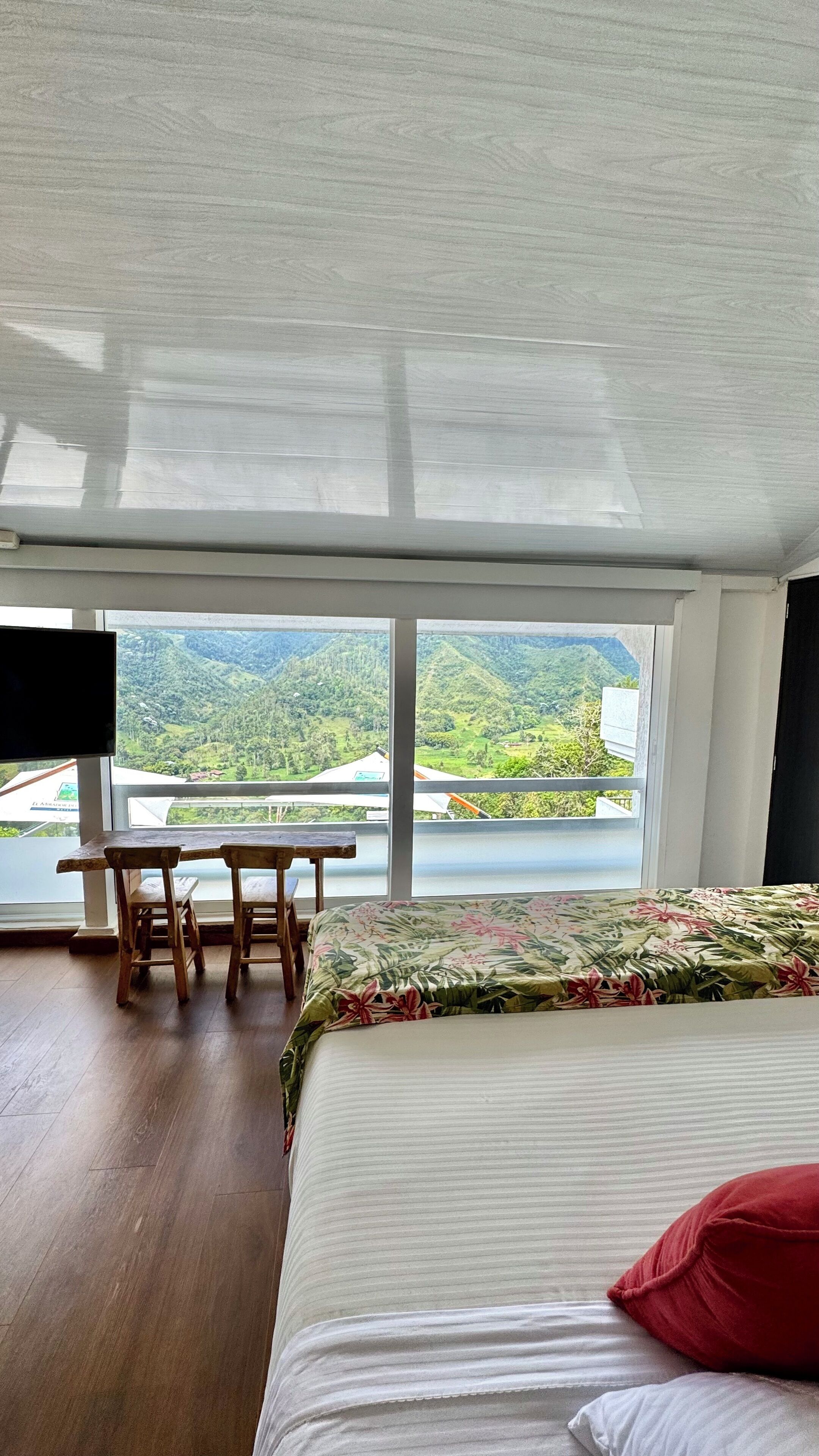 Un dormitorio con un ventanal grande, una cama, un escritorio con dos sillas y vistas a un paisaje montañoso.