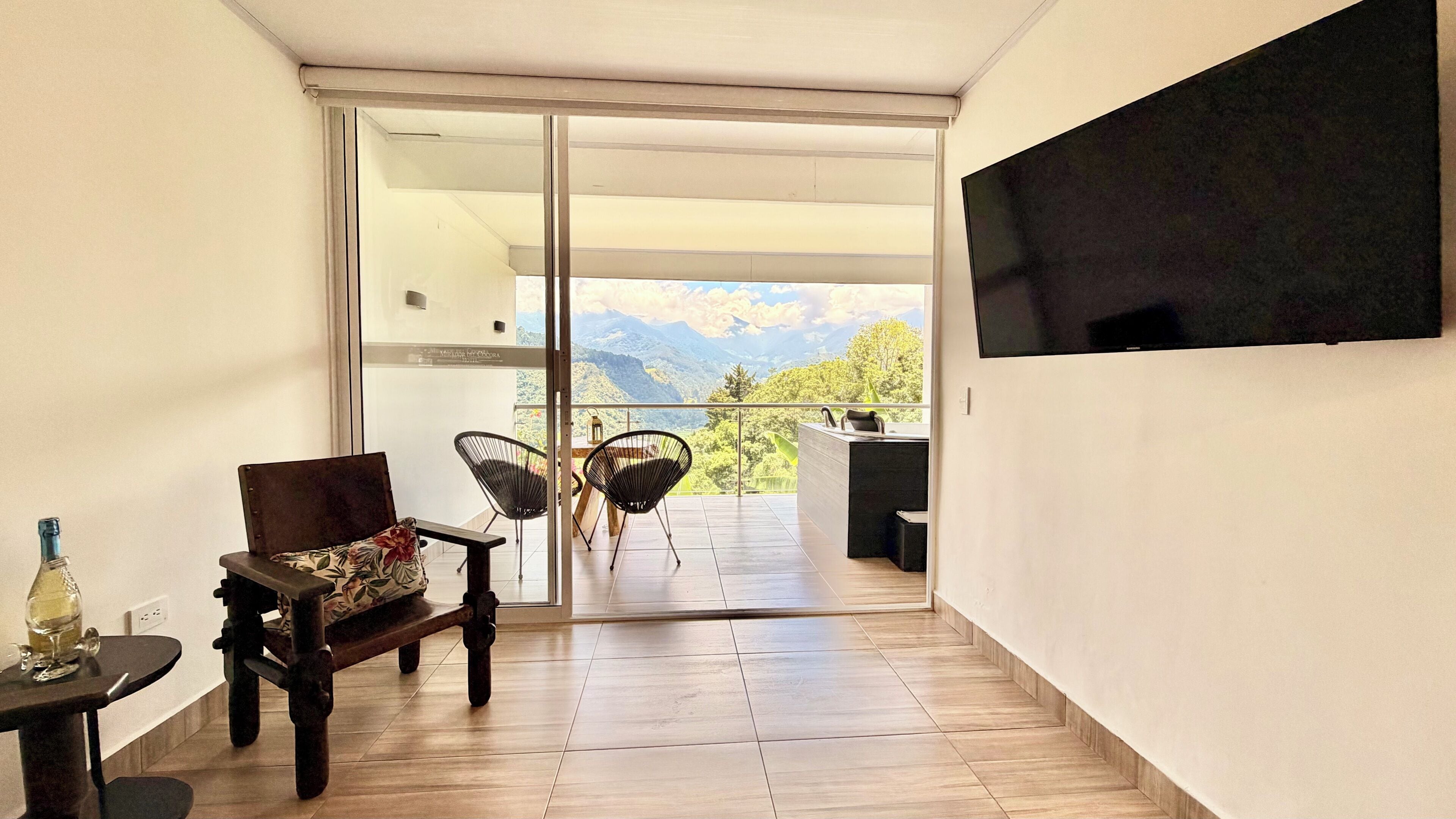 Un intérieur moderne avec un plancher en bois, un canapé, un fauteuil, une petite table avec une bouteille et un téléviseur fixé au mur.