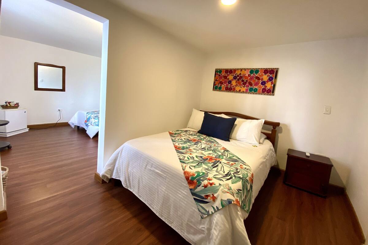A hotel room with a bed, a small desk, a chair, a mirror, and a colorful artwork on the wall.