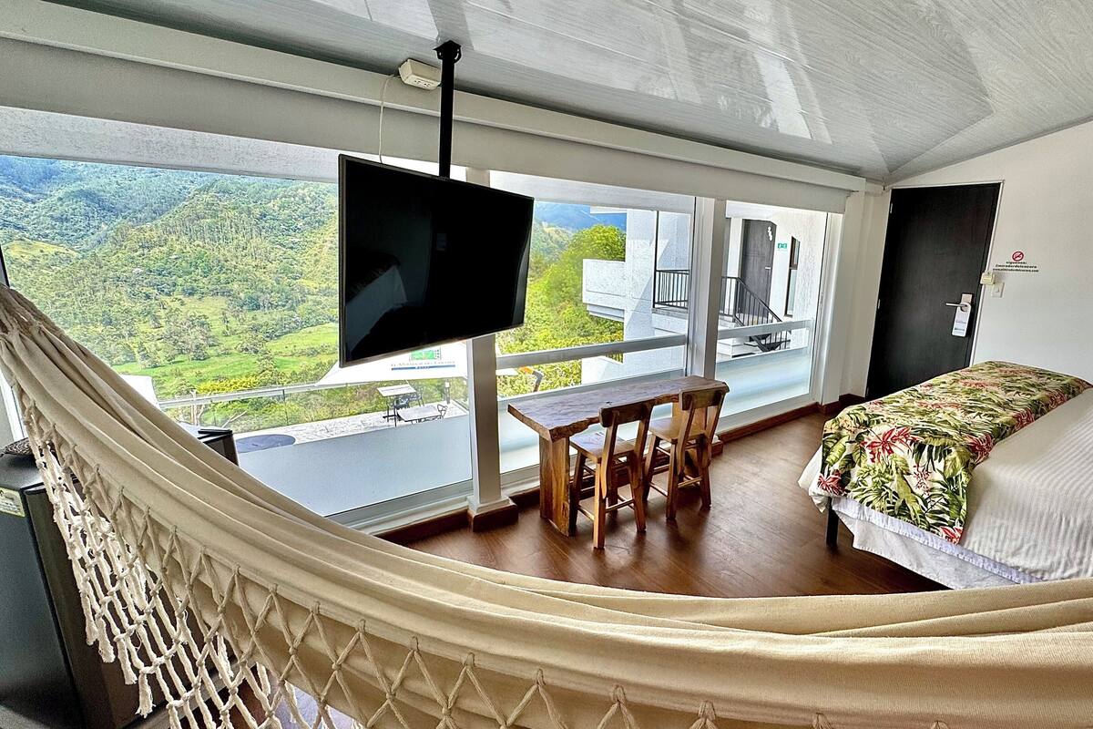 A room with a bed, a TV, a dining table, and a view of a green landscape.