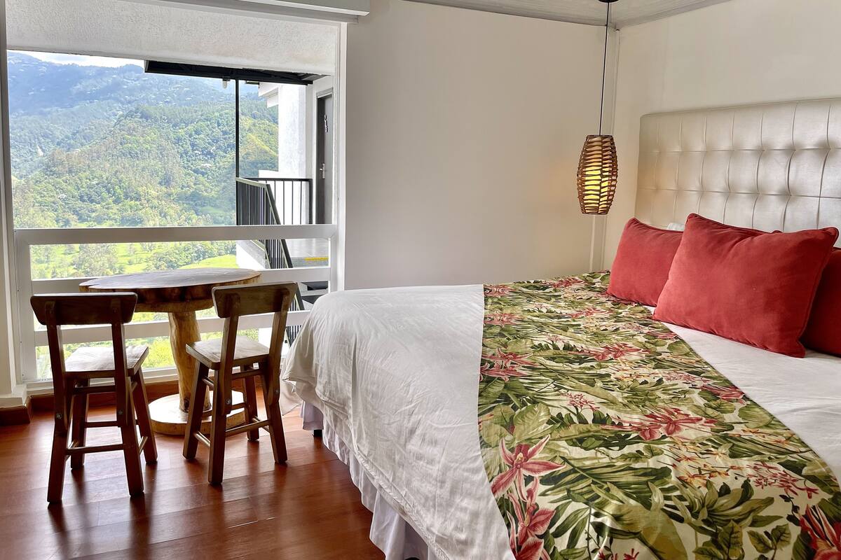 A bedroom with a bed, a window with a view of a forest, a table with chairs, and a hanging light fixture.