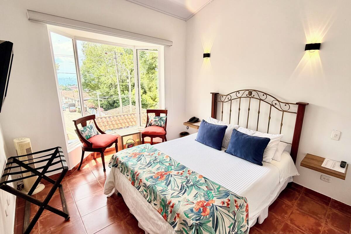 A bedroom with a bed, two chairs, a small table, a heater, and a window with a view of greenery.
