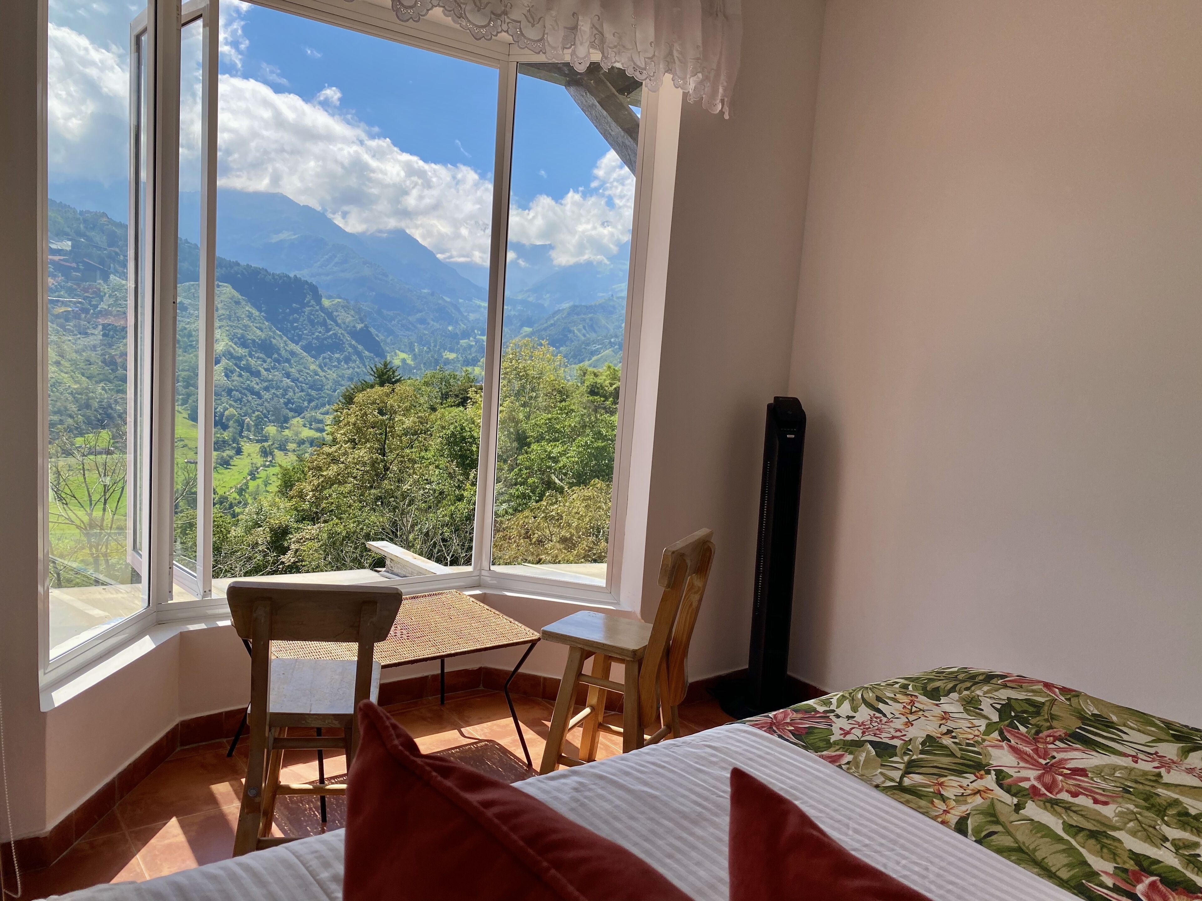 A room with a bed, a table, and a chair, with a large window offering a mountain view.