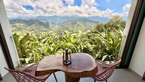 Un balcón con mesa y sillas de madera, con vistas a un paisaje exuberante con montañas y vegetación densa.