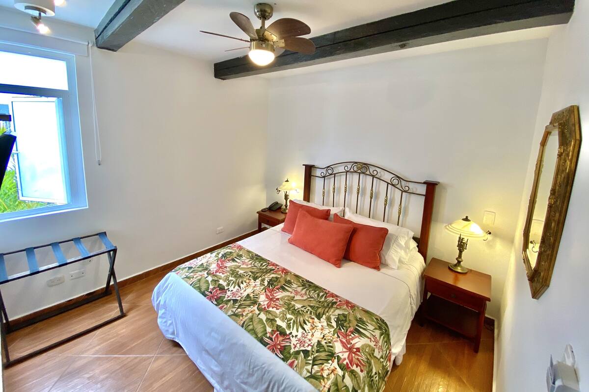 A bedroom with a bed, a ceiling fan, a nightstand with a lamp, a wooden headboard, a window, and a mirror on the wall.