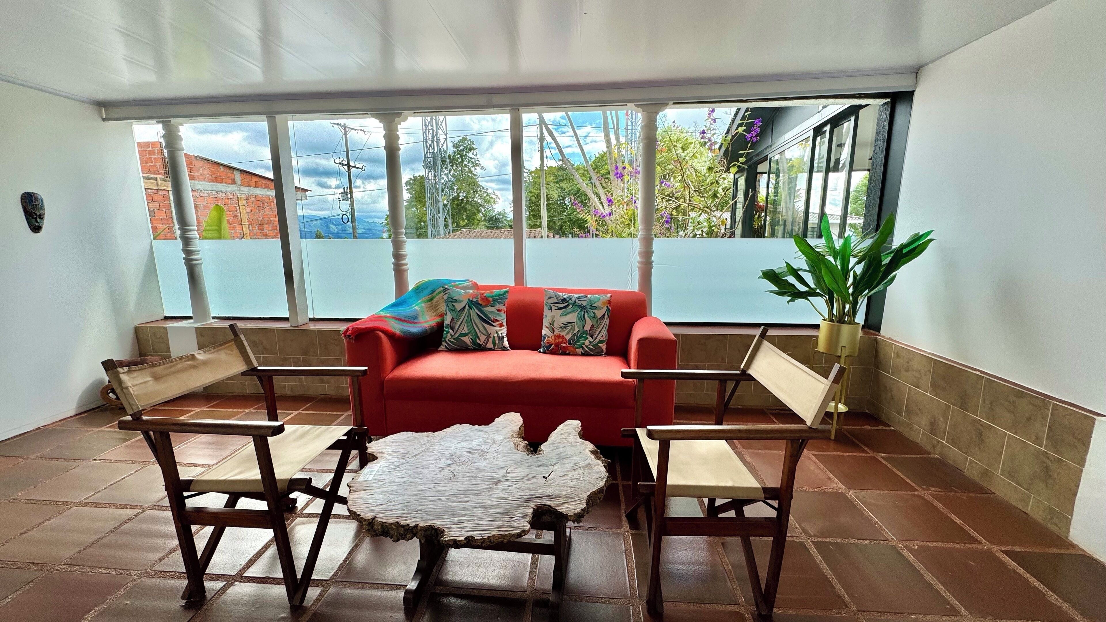 A room with a red sofa, two wooden chairs, a coffee table with a natural wood top, and a large window with a view of greenery and buildings.