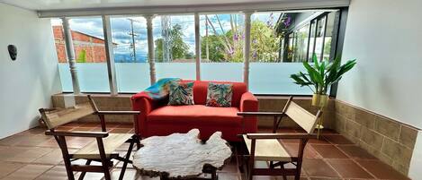 A room with a red sofa, two wooden chairs, a coffee table with a natural wood top, and a large window with a view of greenery and buildings.