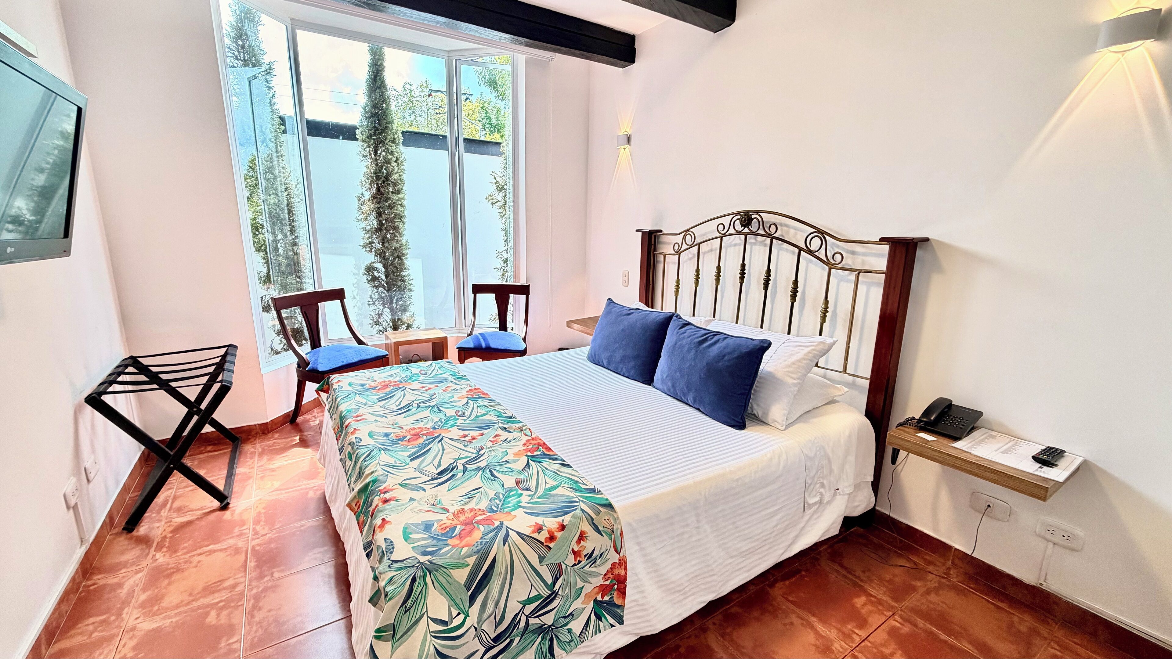 A bedroom with a bed, two chairs, a small table, a window with a view of trees, and a wall-mounted phone.