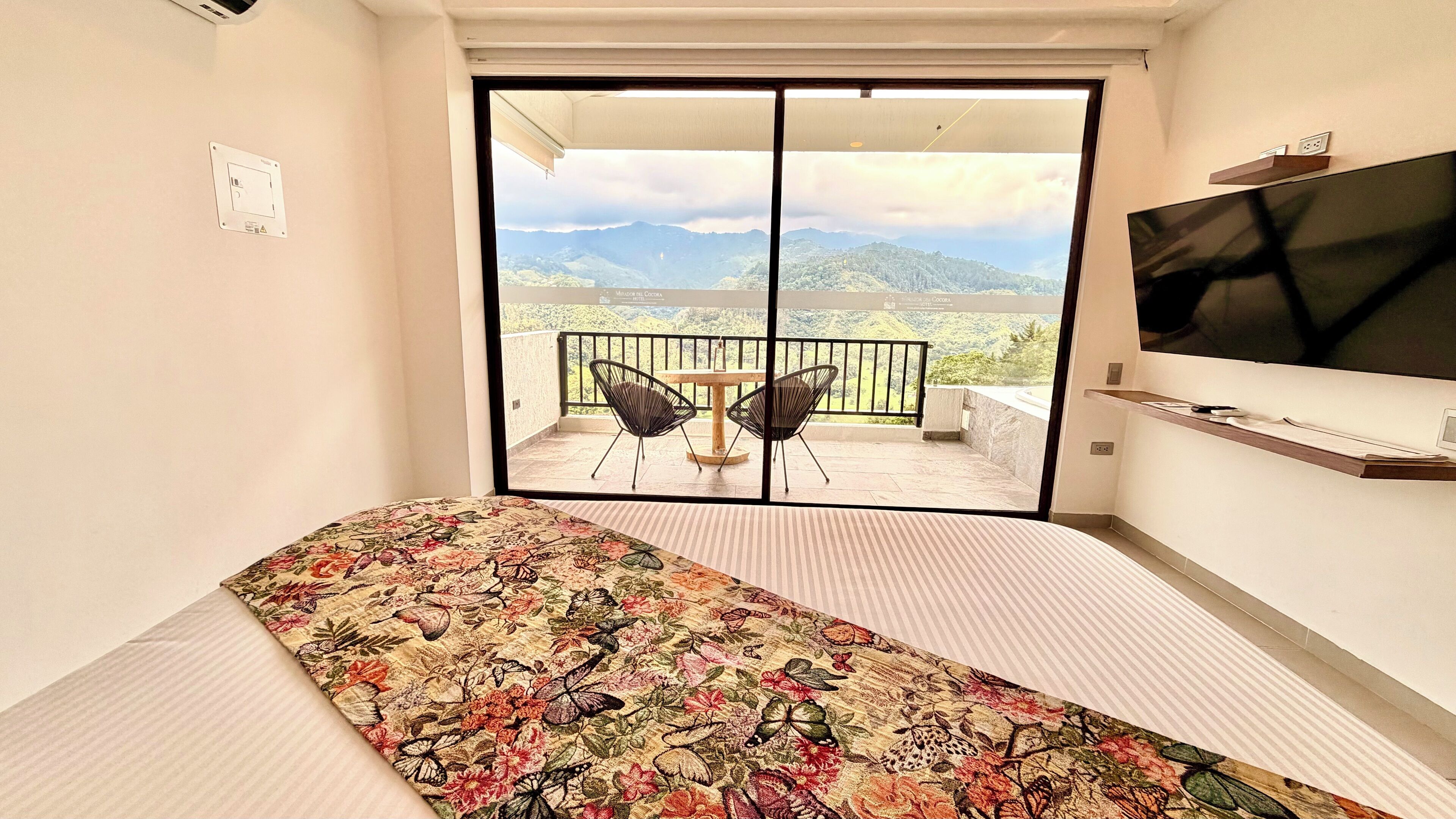 Un dormitorio con cama, televisión, balcón con sillas y vistas a las montañas.