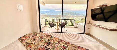 Un dormitorio con cama, televisión, balcón con sillas y vistas a las montañas.