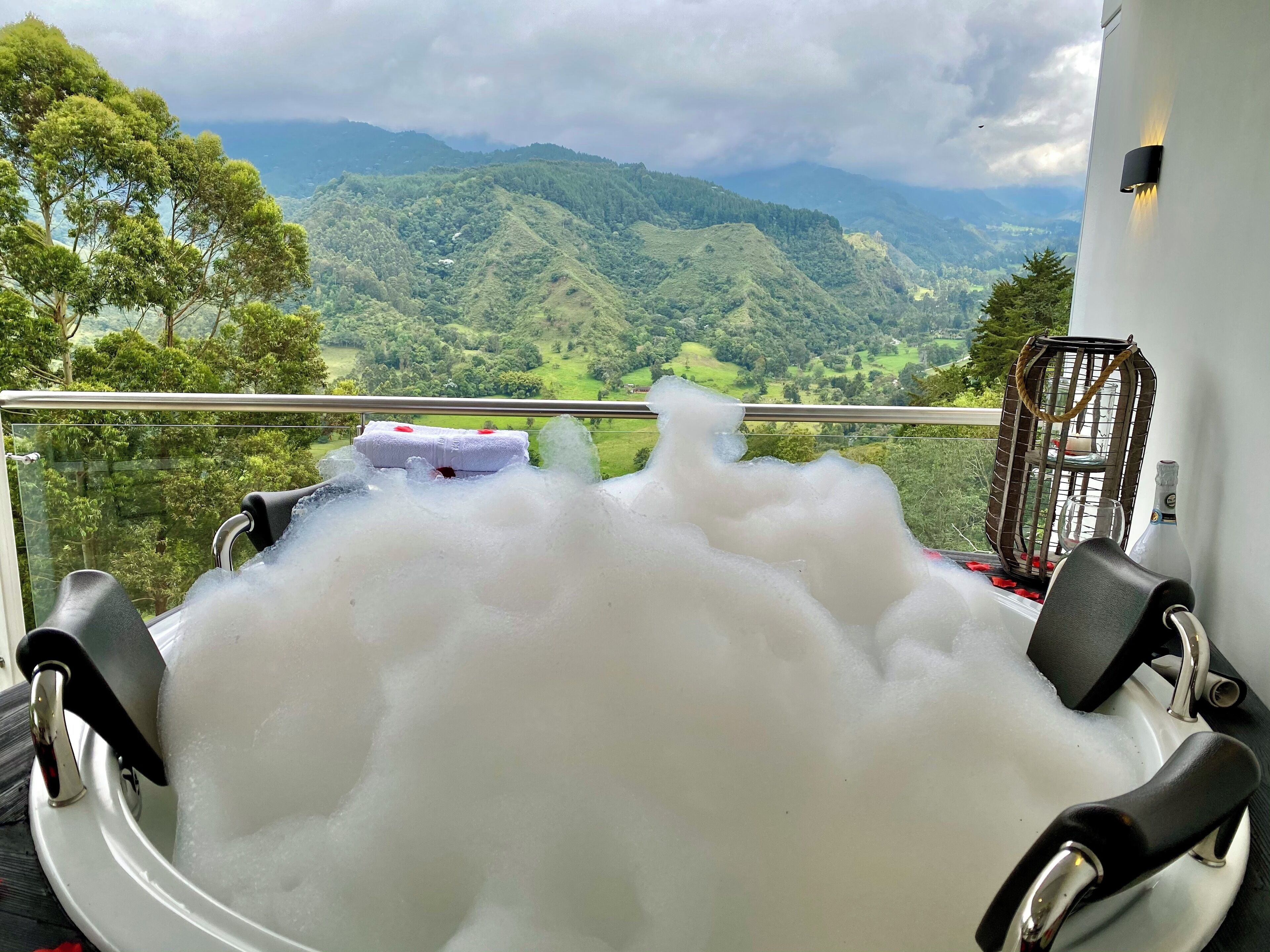 Una bañera llena de burbujas, una toalla y una botella de champán, con una vista panorámica de montañas al fondo.