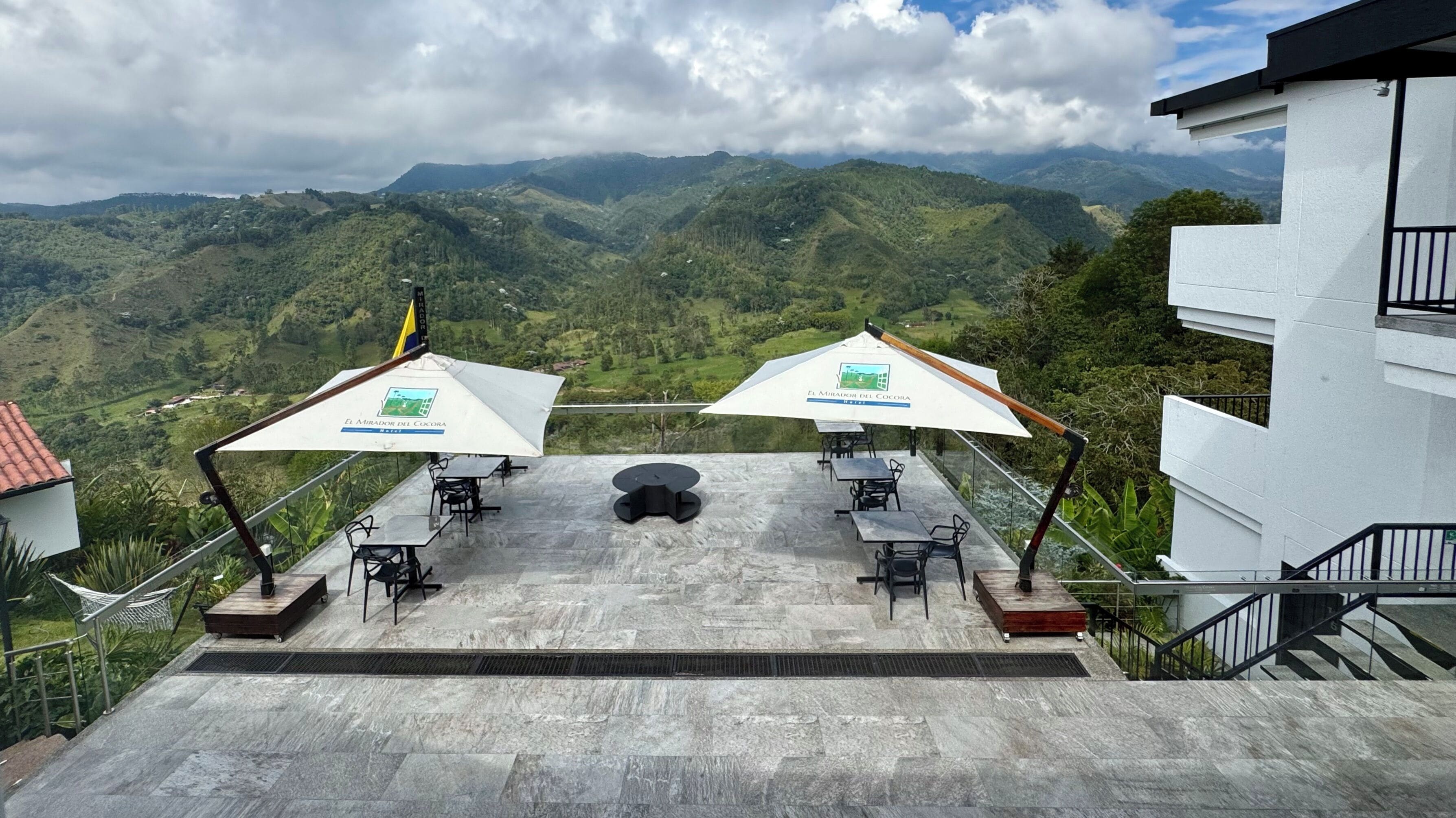A terrace with two tables and umbrellas overlooking a mountainous landscape.
