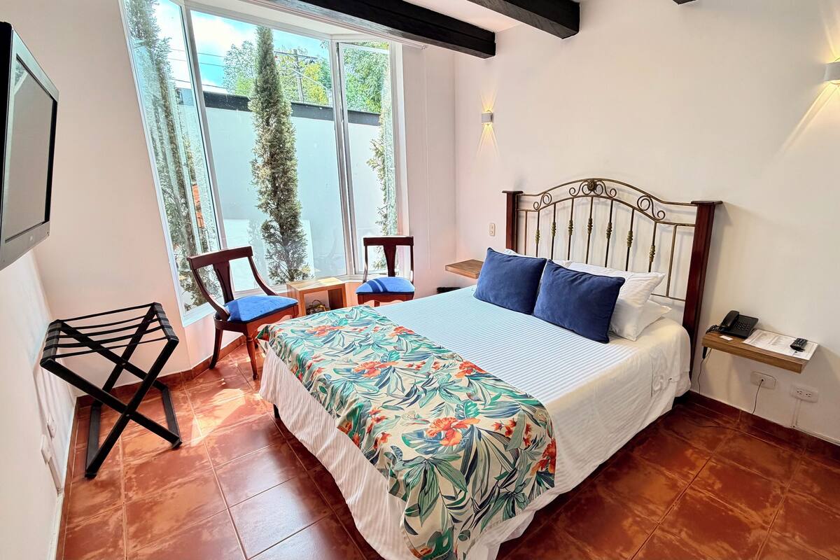 A bedroom with a bed, two chairs, a desk, and a window with a view of trees.