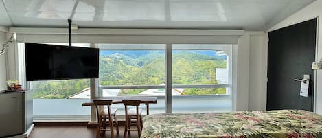 A bedroom with a bed, a TV, a desk with chairs, and a large window offering a view of green hills.