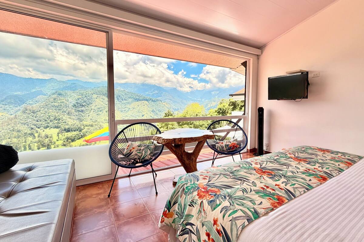 A bedroom with a bed, a sofa, a small table, two chairs, a TV, and a balcony with a mountain view.